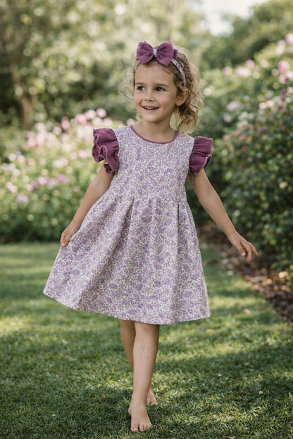 A lifestyle photograph of a young girl with blonde hair walking through a green garden. She is wearing the Larisa dress, which features a light purple paisley print on the body and contrasting deep purple waffle-knit ruffled sleeves. She is also wearing a matching purple waffle bow headband.