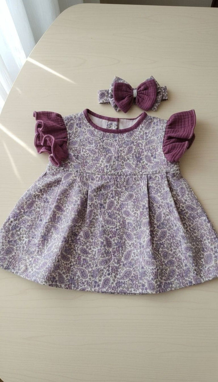 A flat lay photograph of the Larisa baby dress and matching headband. The dress features a lavender and purple paisley print body with plum-colored ruffled muslin sleeves and a matching plum neckline trim. The coordinating headband has a large plum muslin bow with a paisley center, all displayed on a light-colored wooden surface.