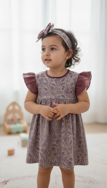 A lifestyle photograph of a young girl with dark curly hair wearing the Larisa dress. The dress features an intricate purple and lavender paisley print with vibrant plum-colored ruffled muslin sleeves and a matching plum crew neckline. She is also wearing a coordinating paisley headband with a plum bow. She is standing and looking off-camera in a bright, airy room with soft-focus wooden toys and a window in the background.