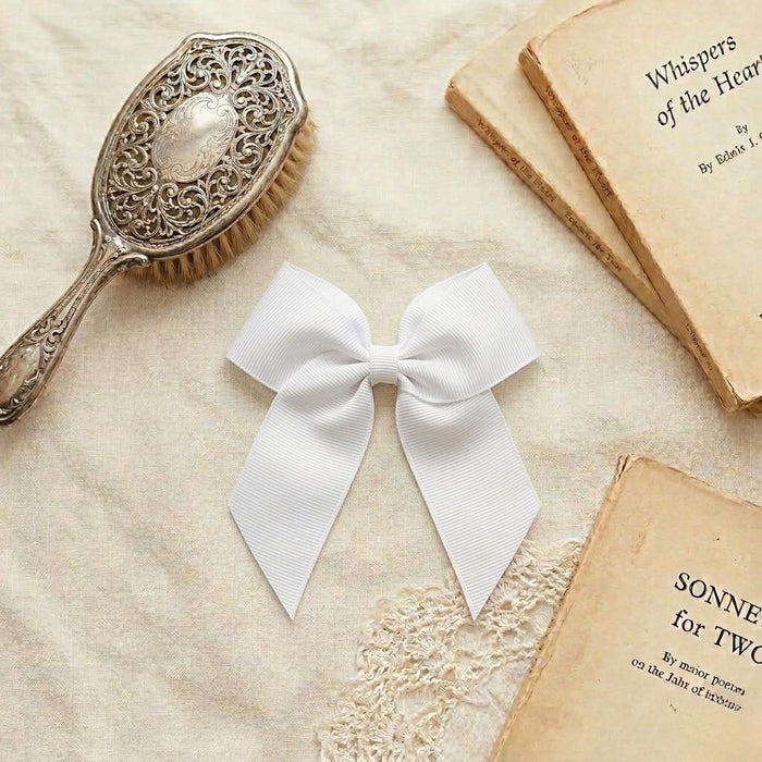 A flat lay photograph of a crisp white grosgrain ribbon hair bow with long tails. The bow is centered on a neutral linen fabric, surrounded by vintage aesthetic props including an ornate silver hairbrush and antique books.