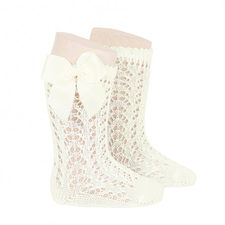 A studio product image of a pair of ivory white knee-high baby socks featuring an intricate, floral-inspired pointelle lace knit. The socks are finished with a scalloped top edge and a large, coordinating ivory satin bow (lazo) on the side.