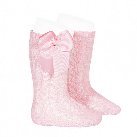 A studio product image of a pair of light pink knee-high baby socks featuring an intricate pointelle lace knit pattern. The socks are designed with a scalloped top edge and a large, coordinating pink grosgrain ribbon bow (lazo) stitched to the side.