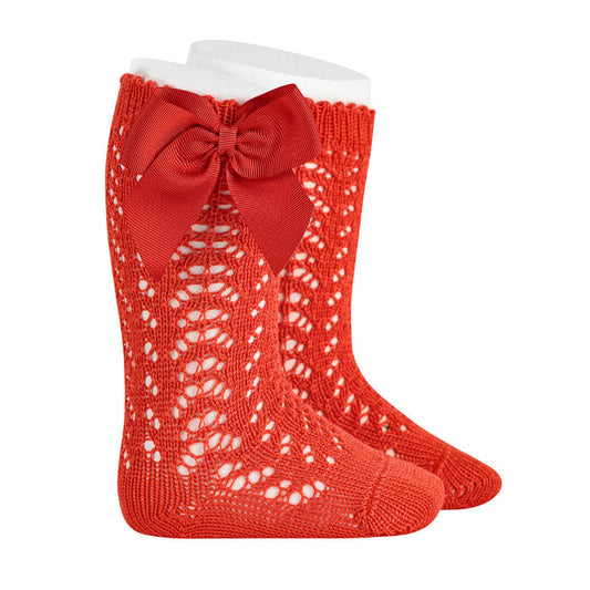 A pair of bright red knee-high baby socks featuring an intricate open-work chevron knit pattern. Each sock is adorned with a large, matching red grosgrain ribbon bow at the outer cuff. The socks are displayed on white forms against a plain white background.