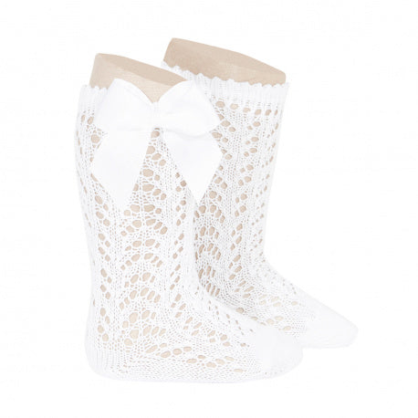 A studio product image of a pair of bright white knee-high baby socks made from a delicate pointelle lace knit pattern. The socks feature a large, matching white grosgrain ribbon bow (lazo) on the side, displayed on light-colored forms against a plain white background.