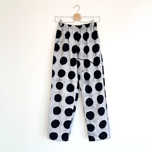 Full front view of Lazy Pants in Indigo Dots Batiki cotton, hanging on a clip hanger against a white wall, showing elastic waistband, bold hand-printed oversized black polka dots on a grey-white ground, and relaxed wide-leg silhouette