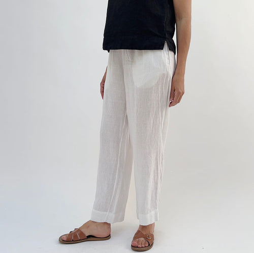 A person standing against a white background, wearing white linen pants and a black top, with tan sandals.