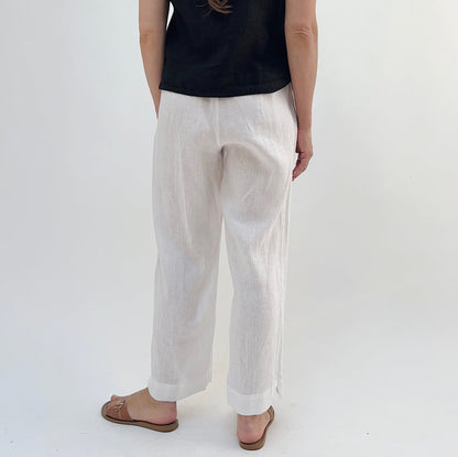 A person standing against a white background, wearing white linen pants and a black top, with tan sandals, viewed from the back