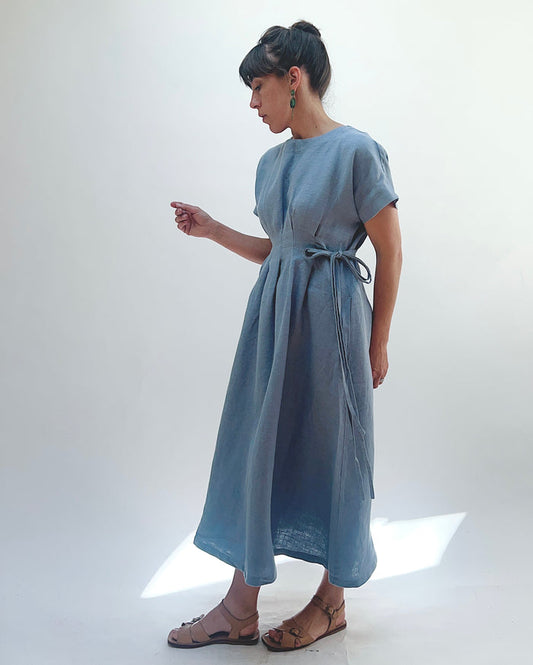 Woman with dark hair in a bun wearing a light blue linen dress and green dangle earrings standing against a white background.