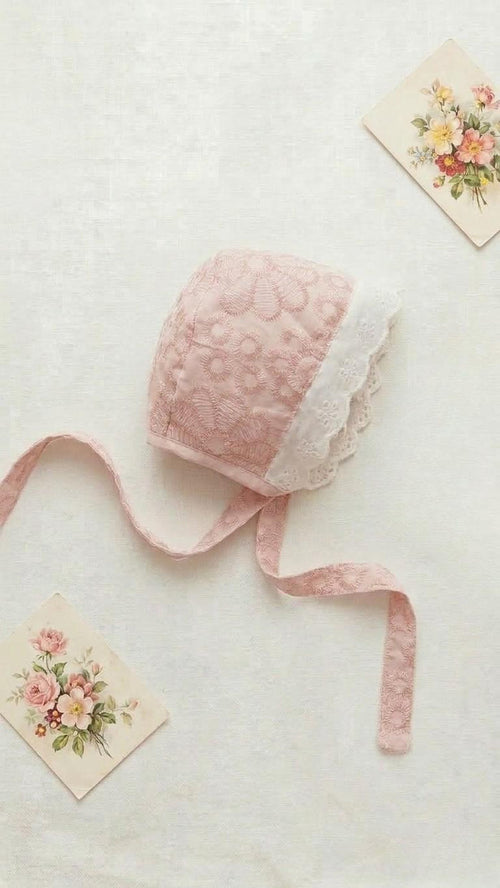 A flat lay of the Lola baby bonnet in a soft dusty pink, featuring intricate floral lace embroidery across the body. The bonnet is trimmed with a delicate white scalloped lace eyelet edge and has matching pink lace ties. It is styled on a neutral linen background with two vintage floral postcards.