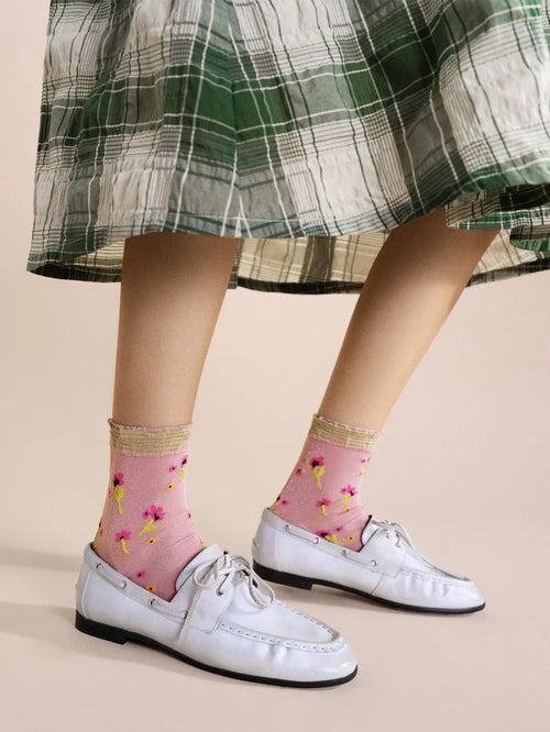 Lumiesta Floral Short Crew socks in Pink — sheer pink nylon short crew with allover floral motif and striped ruffle cuff, worn with white leather boat shoes under a green plaid midi skirt on a cream background