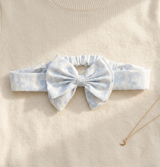 A flat lay photograph of the Malaga baby headband, featuring a soft white fabric with a light blue damask-style swirl pattern. The headband includes an elasticated back for comfort and a large matching fabric bow at the centre. It is styled on a cream-colored knitted sweater background next to a delicate gold moon pendant necklace.