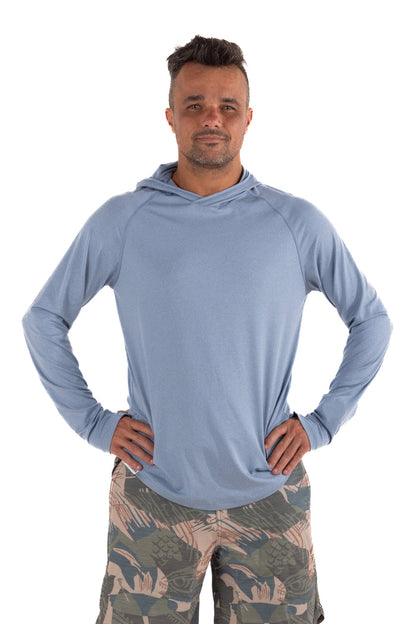 Male model wearing the Waihana Mariner Blue Hooded Long Sleeve sun shirt in light blue with raglan sleeves, paired with tropical-print board shorts, on a white background.