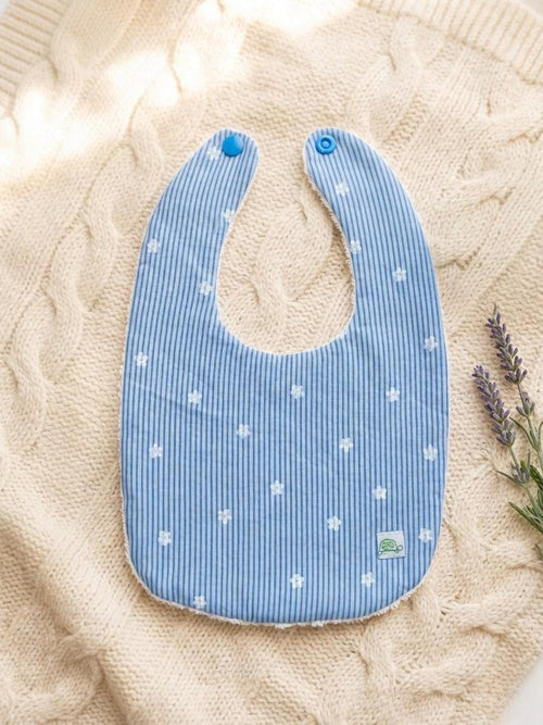 A flat lay of the Menorca baby bib featuring vertical blue and white stripes with tiny white flower motifs scattered across the fabric. The bib has blue snap closures and a small green turtle logo tag, styled on a cream textured cable-knit blanket with a sprig of lavender on the side.