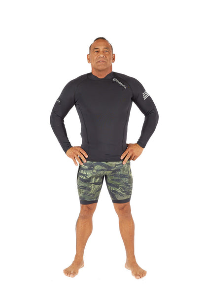 Male model wearing a black Waihana long-sleeve surf rashguard and olive camo Tropicam Tech Jammers wetsuit shorts, standing hands-on-hips against a white background.
