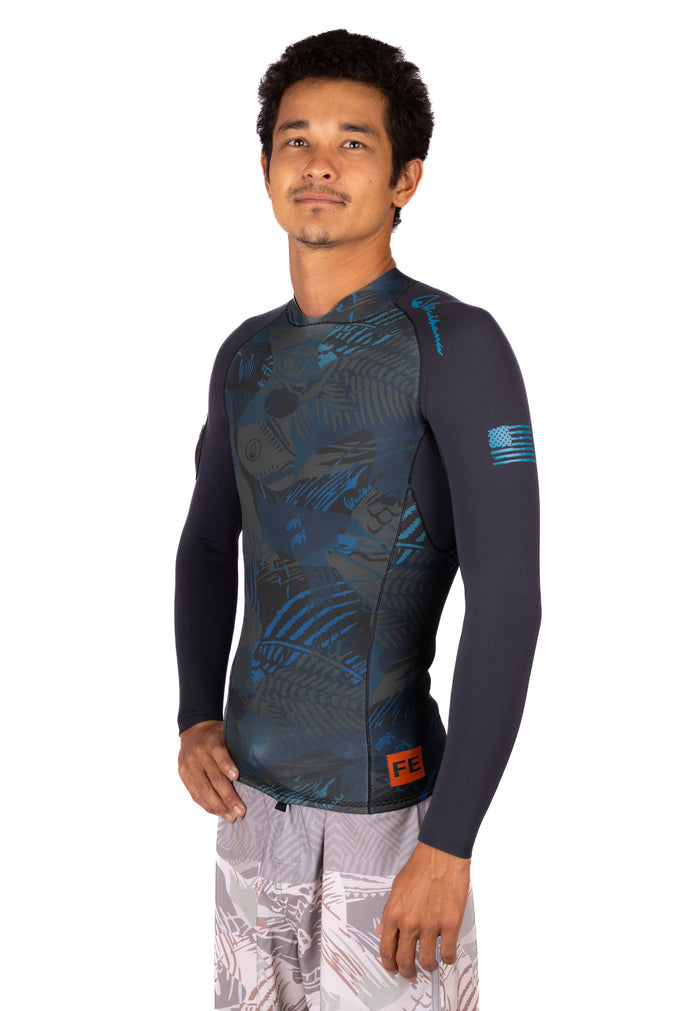 Male model wearing the Waihana Men's Field Ethos Rear-zip Surf Rashguard in FE Blue, a long-sleeve top with a dark tropical camo print front panel, navy sleeves, and an American flag detail on the left arm.