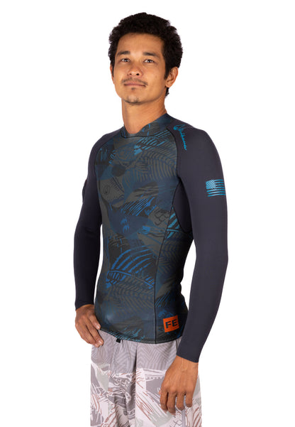 Male model wearing the Waihana Men's Field Ethos Rear-zip Surf Rashguard in FE Blue, a long-sleeve top with a dark tropical camo print front panel, navy sleeves, and an American flag detail on the left arm.