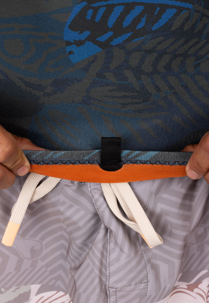 Close-up of a man lifting the hem of a Waihana Men's Field Ethos blue patterned surf rashguard to show the orange-trimmed waistband and cream drawstring of grey patterned board shorts underneath.