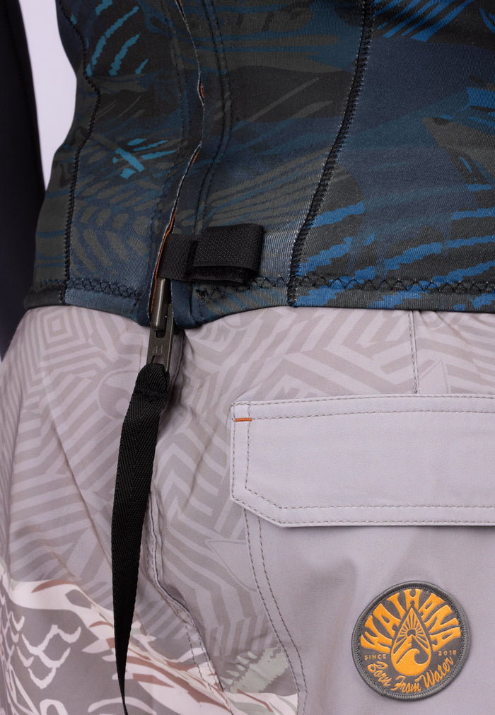 Close-up of the waist area of a Waihana Men's Field Ethos Rear-zip Surf Top in blue camo pattern, showing the zipper pull clip and Waihana branded logo patch on grey patterned board shorts.