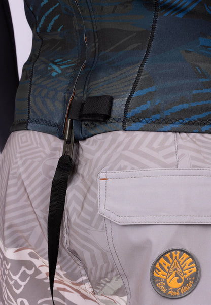 Close-up of the waist area of a Waihana Men's Field Ethos Rear-zip Surf Top in blue camo pattern, showing the zipper pull clip and Waihana branded logo patch on grey patterned board shorts.