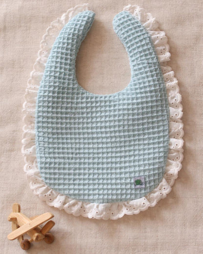 A flat lay photograph of a light mint green baby bib made from textured waffle-weave fabric. The bib is detailed with a white ruffled eyelet lace trim along the edges and features a small green turtle brand logo. It is styled on a neutral linen surface next to a small wooden toy airplane.