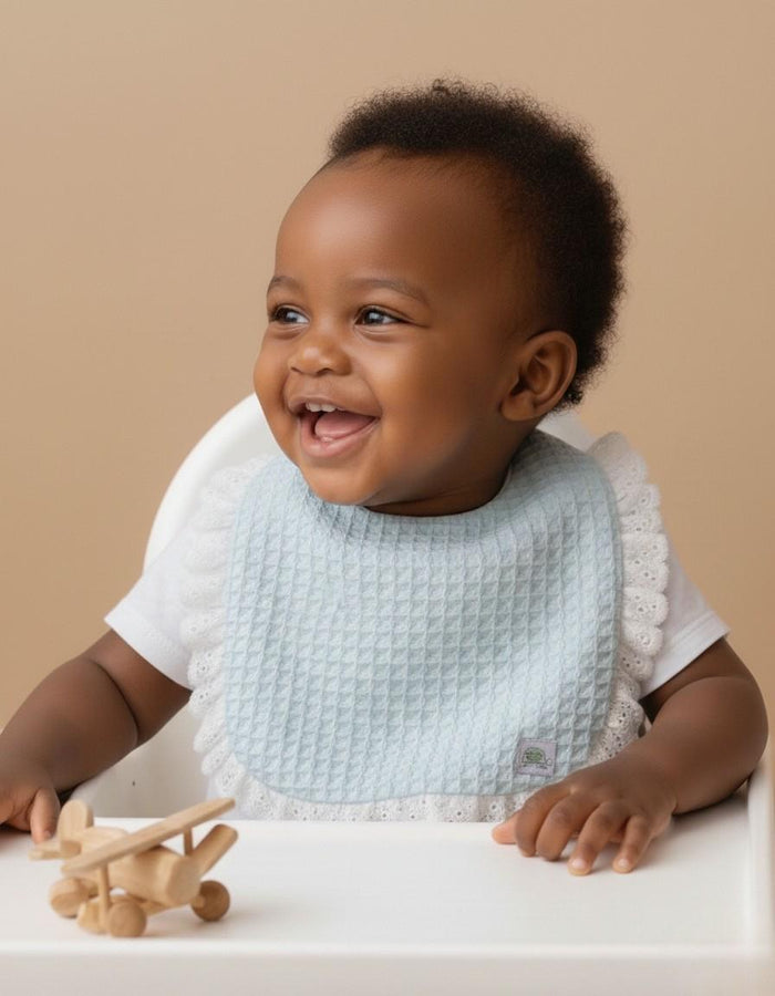 A lifestyle photograph of a laughing baby with dark hair sitting in a white highchair. The baby is wearing a light mint waffle-weave bib with white ruffled eyelet lace trim and a small brand label. A small wooden toy airplane sits on the highchair tray in the foreground against a warm, neutral tan background.