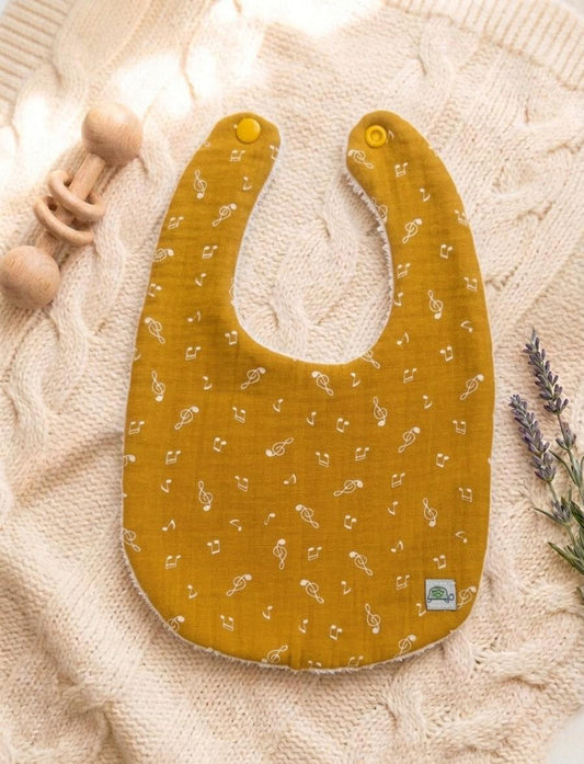 A flat lay of a mustard yellow baby bib made from soft muslin cotton, featuring a delicate white pattern of musical notes and treble clefs. The bib has two coordinating yellow snap closures and is displayed on a pink cable-knit sweater next to a wooden rattle and lavender sprigs.