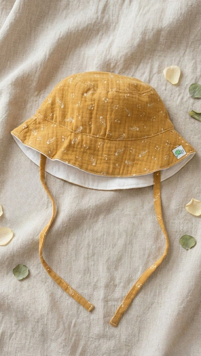 A flat lay photograph of a baby bucket hat made from mustard yellow muslin fabric with a white music note print. The hat features matching fabric chin straps and a small green turtle brand label on the brim. It is styled on a neutral linen backdrop with scattered white flower petals and green leaves.