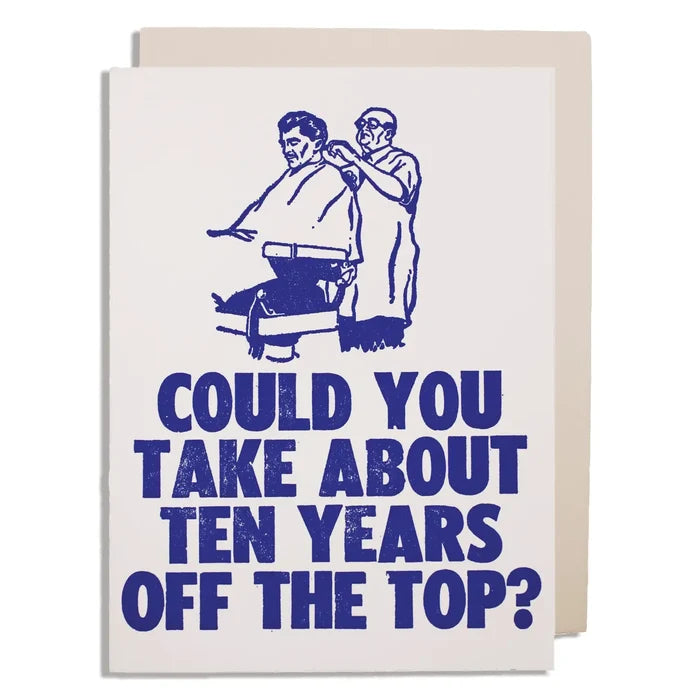 A square greeting card with a light cream background and a blue graphic of a barber and customer, with the text 'COULD YOU TAKE ABOUT TEN YEARS OFF THE TOP?' in large blue letters. A second cream card or envelope is partially visible behind it on a white background.