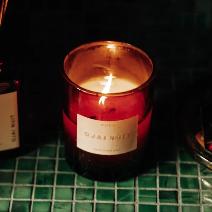Candle in a copper container labeled 'Ojai Nuit' on a green grid surface.