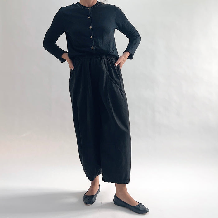 Full front view of model wearing Eleven Stitch black one pocket pant with hands on hips, showing elastic waistband, wide cropped leg, curved side pocket, gathered elastic hem, and black color, styled with black long sleeve button front top and black ballet flats