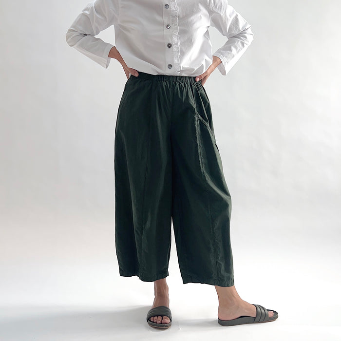 Full front view of model wearing Eleven Stitch konbu one pocket pant with hands on hips, showing elastic waistband, wide curved-seam cropped leg, large curved side pocket, and deep forest green color, styled with white long sleeve button front shirt and dark slide sandals