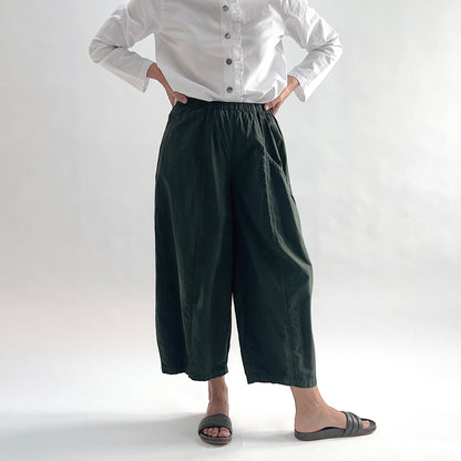 Full front view of model wearing Eleven Stitch konbu one pocket pant with hands on hips, showing elastic waistband, wide curved-seam cropped leg, large curved side pocket, and deep forest green color, styled with white long sleeve button front shirt and dark slide sandals