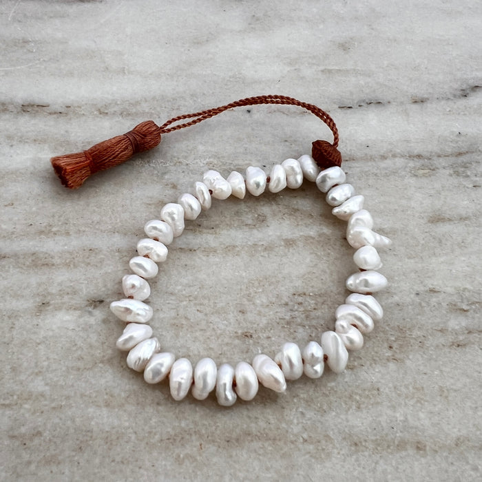 A white pearl tassel bracelet with a brown waxed silk cord and an adjustable tassel closure, displayed on a marble surface.