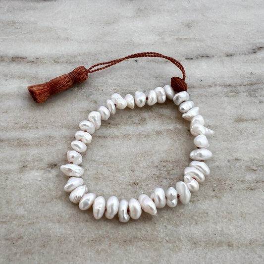 A white pearl tassel bracelet with a brown waxed silk cord and an adjustable tassel closure, displayed on a marble surface.
