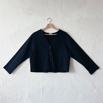 A black, long-sleeved crop cardigan with a button front, displayed on a hanger against a white textured wall.