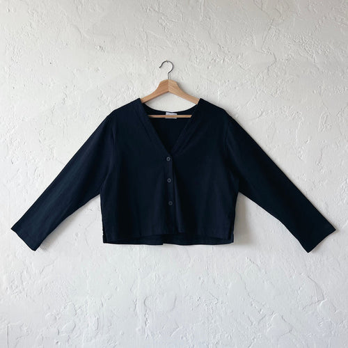 A black, long-sleeved crop cardigan with a button front, displayed on a hanger against a white textured wall.