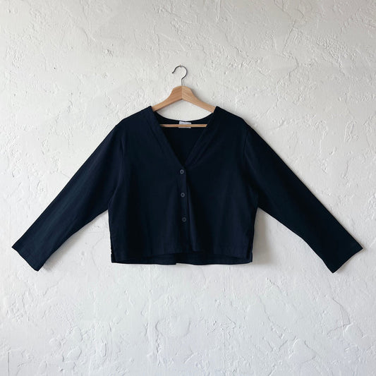 A black, long-sleeved crop cardigan with a button front, displayed on a hanger against a white textured wall.