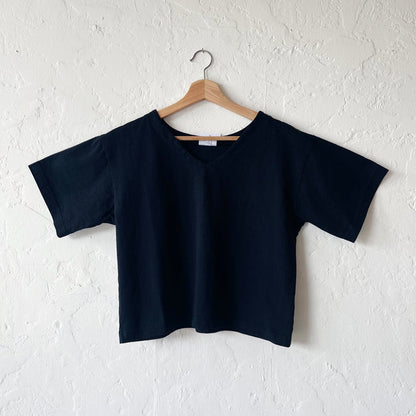 A black, cropped sleeve top with a slight v-neck hanging on a hanger against a white textured wall.