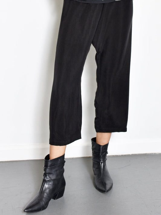 A person wearing black cupro pants and black boots standing against a white background.