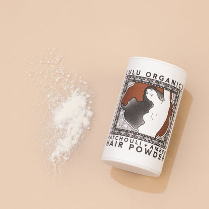 Overhead view of Lulu Organics Patchouli and Amber Hair Powder Shampoo container lying on its side next to a small pile of white powder on a tan background