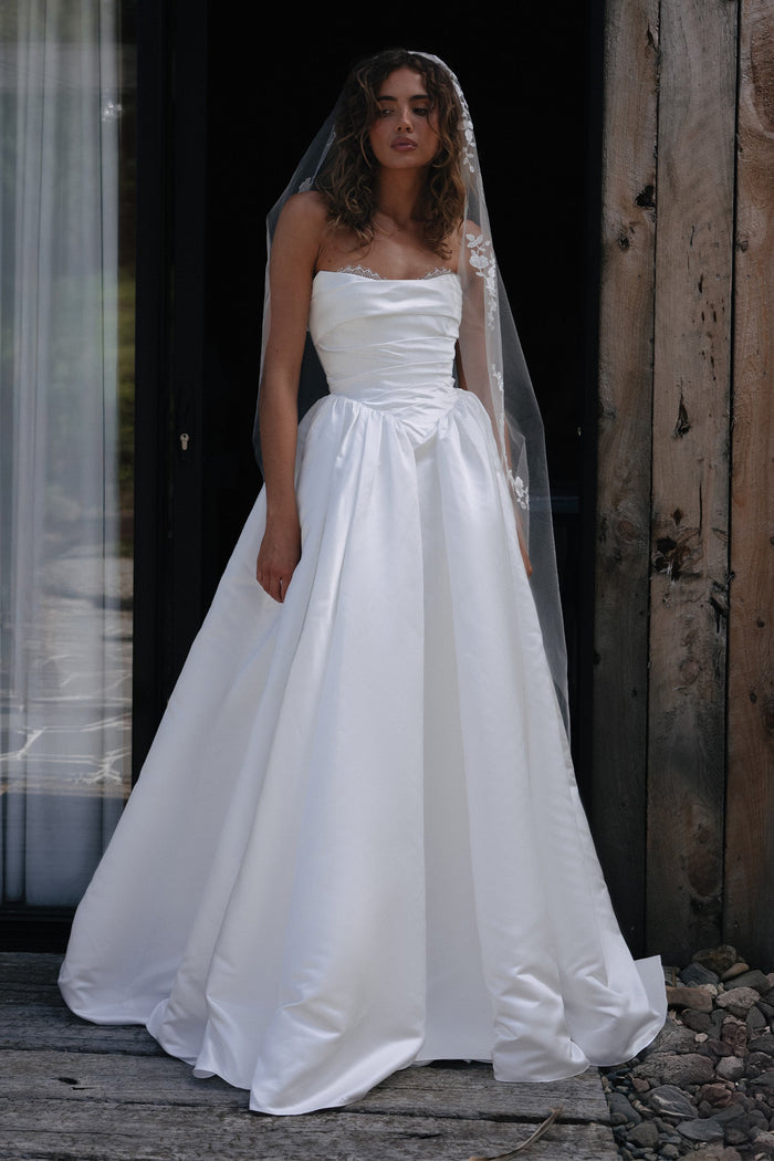 Woman wearing a white wedding dress with a veil standing against a rustic wooden wall.