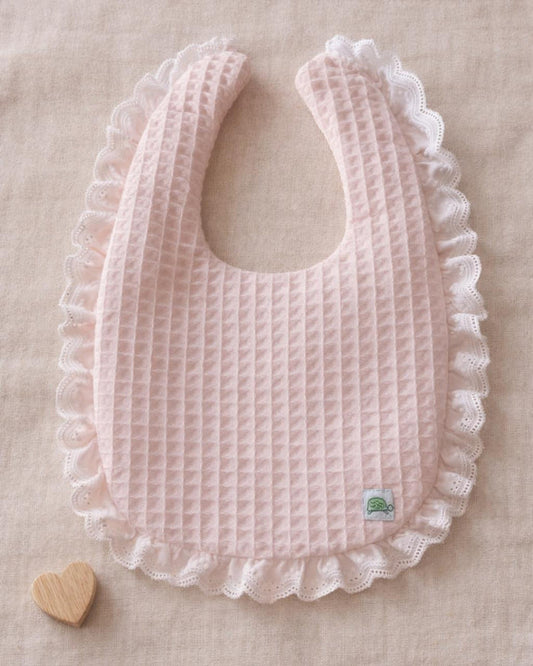 A flat lay photograph of a light pink baby bib made from textured waffle-weave fabric. The bib features a delicate, white ruffled eyelet lace trim around the entire outer edge and a small green turtle brand label in the lower right corner. It is styled on a neutral linen backdrop next to a small wooden heart.