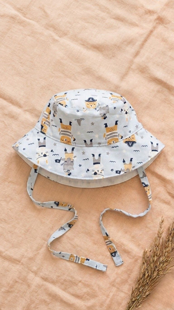 A flat lay photograph of a baby bucket hat featuring a light blue fabric with a pirate-themed print, including anchors, treasure maps, and stars. The hat has matching fabric chin straps and is styled on a neutral linen backdrop with a sprig of dried wheat in the lower right corner.