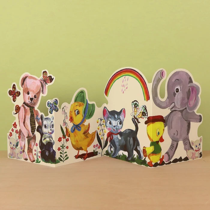 Plushie Party die-cut concertina card by Nathalie Lété, partially open showing a pink teddy bear with a tie and a skunk alongside a black cat, yellow chick, and elephant, with butterflies and a rainbow, against a green background