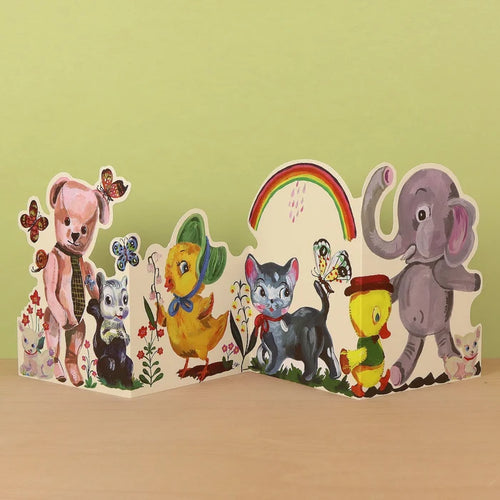 Plushie Party die-cut concertina card by Nathalie Lété, partially open showing a pink teddy bear with a tie and a skunk alongside a black cat, yellow chick, and elephant, with butterflies and a rainbow, against a green background