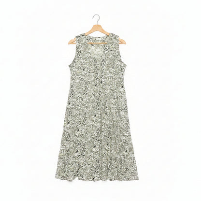 A sleeveless A-line dress with a dense green floral pattern on a white base, hanging on a wooden hanger on a dark concrete floor.