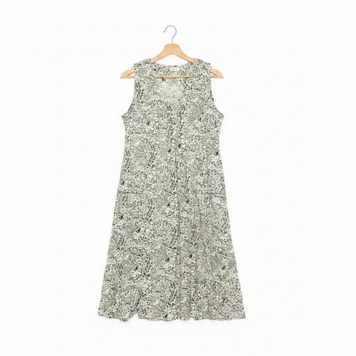 A sleeveless A-line dress with a dense green floral pattern on a white base, hanging on a wooden hanger on a dark concrete floor.