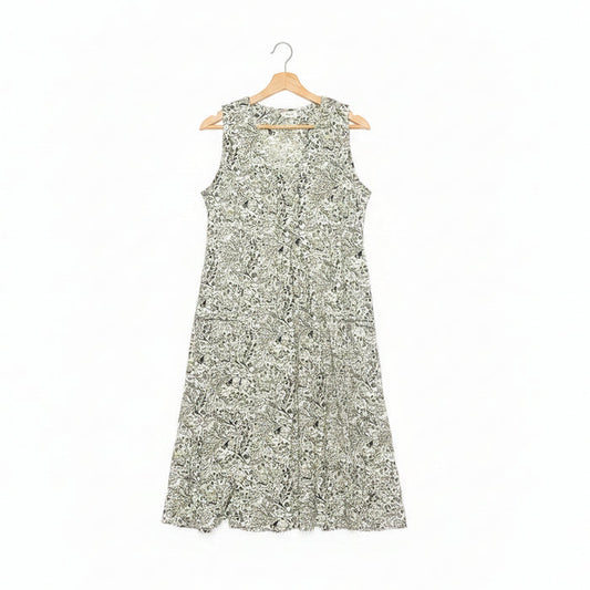 A sleeveless A-line dress with a dense green floral pattern on a white base, hanging on a wooden hanger on a dark concrete floor.