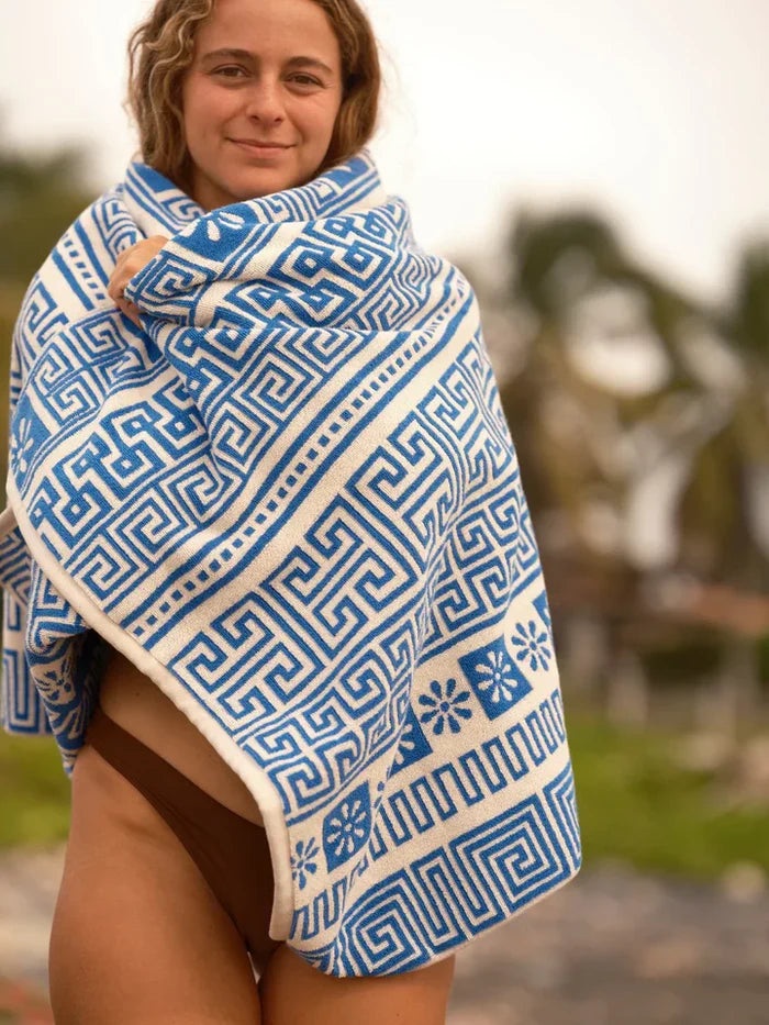 Female model wrapped in the Mollusk Surf Shop Poseidon Towel, smiling outdoors, showing full blue and white jacquard Greek key pattern with floral motifs and cream border
