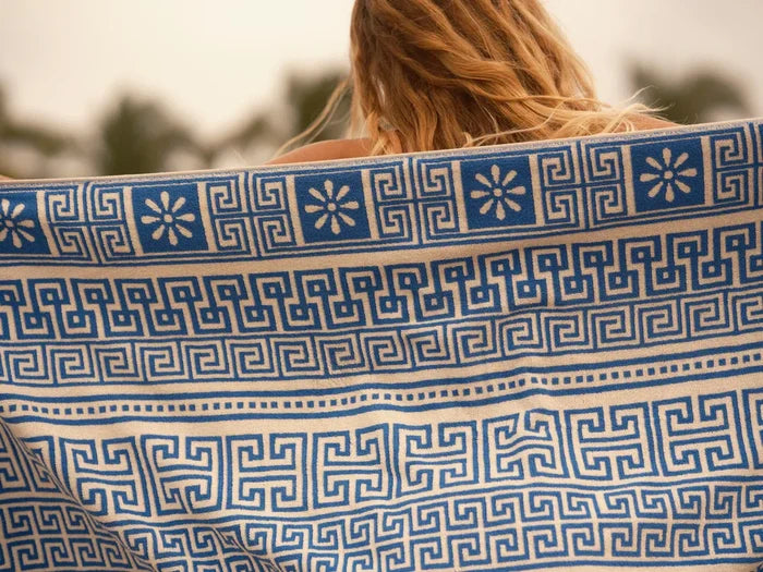 Close-up detail of the Mollusk Surf Shop Poseidon Towel held up outdoors, showing blue and natural white jacquard weave with Greek key meander pattern and daisy floral motifs